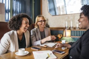 Três mulheres conversando sobre como fazer brainstorming em volta de uma mesa com xícaras, papéis e canetas.