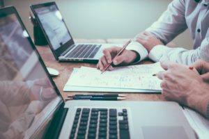 Mesa com dois notebooks, canetas e papéis, com pessoas conversando sobre consultoria empresarial.