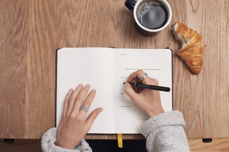 Mulher fazendo uma pauta de reunião em uma mesa com uma xícara de café e um croissant